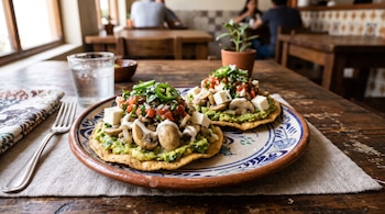 Dos tostadas con aguacate, champiñones, queso blanco y salsa en un plato decorado, junto a un tenedor y un vaso con agua sobre una mesa.