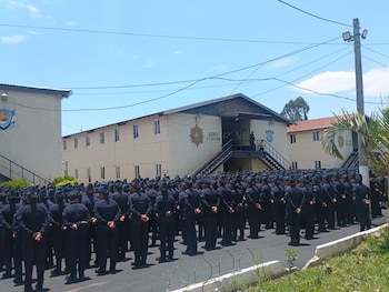 Vista trasera de personal uniformado de oscuro en formación frente a un edificio beige con el letrero 'QUINTA ESTACION' bajo cielo azul. Poste eléctrico y palmeras