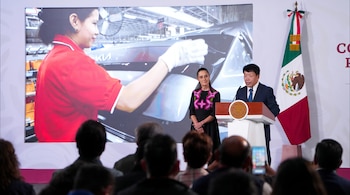 Un hombre habla en un podio junto a una mujer, mientras una pantalla grande proyecta una mujer trabajando en una fábrica automotriz. Se observa una bandera de México.