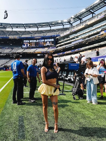 Mujer con gafas de sol, top negro, falda amarilla y micrófono sobre césped. Al fondo, un estadio con pantalla que muestra "FIFA World Cup 2026"