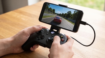 Dos manos sostienen un mando negro acoplado a un teléfono móvil que muestra en pantalla un automóvil rojo sobre una pista de carreras.
