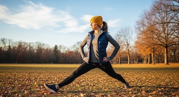 Una mujer vista de cuerpo entero, con gorro amarillo, chaleco azul y mallas negras, se estira con las piernas abiertas en un parque con hojas secas en el suelo.