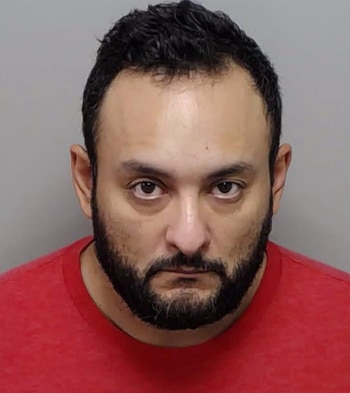Fotografía policial de un hombre adulto con cabello oscuro, barba y camiseta roja sobre un fondo gris