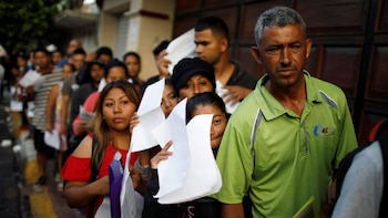 Una larga fila de personas de diversas edades, algunas con papeles blancos, se extiende en un entorno urbano. Un hombre con camisa verde es visible al frente