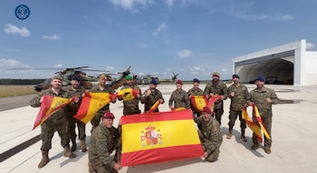 Once militares con uniformes de camuflaje posan al aire libre, sosteniendo banderas de España, con helicópteros y un hangar al fondo