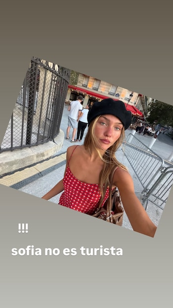 Mujer joven con vestido rojo y boina negra. De fondo, una calle con vallas, edificios y personas. Contiene texto