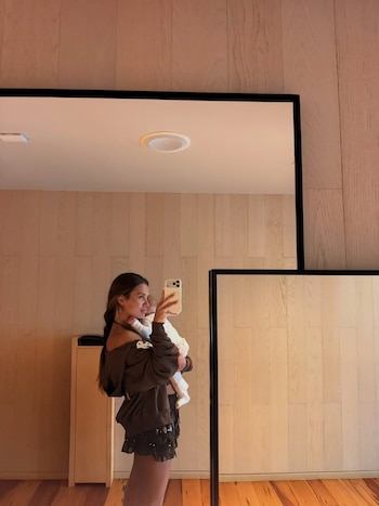Uma mulher segurando um bebê enquanto se olha no espelho e tira uma foto com um celular, em uma sala com paredes e piso de madeira