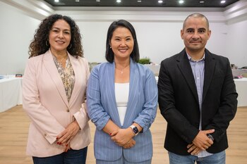 Mujer con cabello rizado y blazer rosa, mujer con cabello liso y blazer azul a rayas, hombre con camisa a rayas y blazer negro, sonriendo de pie en sala