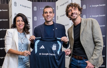 Tres personas sonríen a la cámara, dos hombres y una mujer, sosteniendo una camiseta de fútbol azul oscura con el logo de Google Gemini al frente. Detrás hay un fondo con logos de AFA y Google Gemini