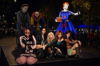Cinco jóvenes sobre una plataforma frente a dos personas disfrazadas y una gran escultura iluminada de catrina en una calle nocturna.