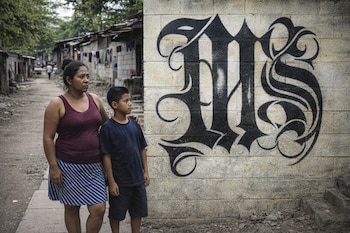 Una mujer y un niño de pie en un camino de tierra. Al fondo se ven casas humildes y a la derecha un muro de concreto con grafiti negro de la letra MS.