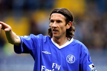 Hombre de cabello castaño y diadema negra con camiseta azul de fútbol del Chelsea FC, escudo del club y logo Umbro, apunta con el brazo izquierdo. Fondo borroso