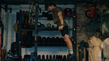 Aldo Kane, un hombre musculoso con barba, realiza fondos en barras paralelas llevando un chaleco lastrado en un gimnasio repleto de equipos