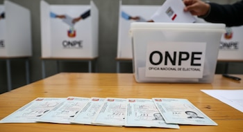 DNI peruanos sobre mesa de madera clara, con una urna de la ONPE y cabinas de votación al fondo; una mano deposita un voto.