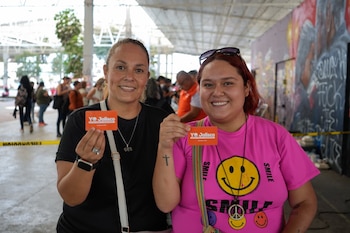 Dos mujeres sonrientes, una con camiseta negra y otra con camiseta rosa, sostienen tarjetas naranja "YO Jalisco" en un entorno público