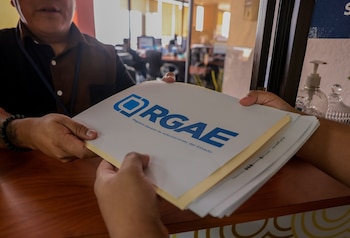 Dos personas intercambian un expediente con la inscripción RGAE en una oficina. Se observa un dispensador de gel y objetos de cristal