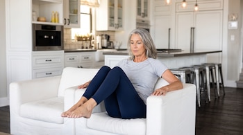 Mujer de cabello canoso con camiseta gris y mallas azules sentada en un sillón blanco elevando las rodillas hacia el pecho.