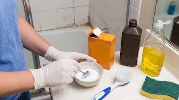 Manos con guantes mezclan una pasta en un tazón junto a una caja de bicarbonato, agua oxigenada, detergente, un cepillo y una esponja.