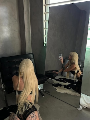 Imagen de La Joaqui arrodillada de espaldas en lencería negra y tatuajes, reflejada en un espejo mientras toma una selfie con su teléfono