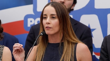 Primer plano de Mara Sedini, una mujer de cabello castaño, hablando en un micrófono. Viste blusa azul marino. Fondo borroso rojo y azul con texto