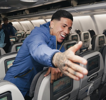 Hombre joven con chaqueta azul, corte de pelo moderno y tatuajes en el brazo, sonriendo y extendiendo una mano dentro de un avión