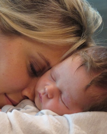 Rostro de mujer rubia con ojos cerrados junto al rostro de un bebé durmiendo, también con ojos cerrados. Ambos recostados sobre tela blanca