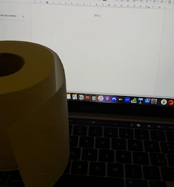 Un rollo de papel higiénico amarillo parcialmente cubriendo el teclado de un portátil, cuya pantalla muestra un documento con el texto "(Fin.)"