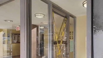 Vista de una puerta de vidrio interior con cinta amarilla de "CAUTION" y "CUIDADO" pegada, bloqueando el paso y reflejando una señal de salida