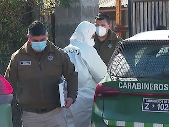 Dos oficiales de Carabineros y un perito forense en traje blanco y mascarillas de pie junto a un vehículo policial verde. Un oficial sostiene un cuaderno