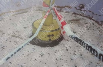Poste metálico amarillo en el centro, atado con cintas de precaución blancas y rojas y cintas blancas con texto negro, sobre suelo irregular