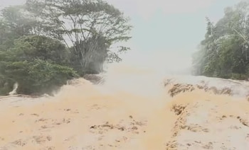 Agua turbia de un río desbordado ocupa el primer plano. Árboles con abundante follaje se observan en las riberas izquierda y derecha. Cielo cubierto