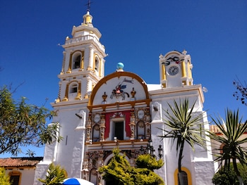 Iglesia blanca con torre de campanario y torre de reloj, decoraciones amarillas y rojas, escultura ecuestre en el frente, cielo azul, árboles y plantas