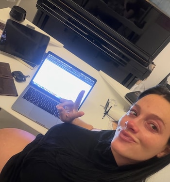 Oriana Sabatini, con cabello oscuro y camiseta negra, sonríe a cámara haciendo un gesto de 'rock and roll' con su mano, sentada frente a un laptop con pantalla brillante y un piano negro al fondo