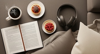 Una taza de café, un libro abierto, dos postres pequeños, unos auriculares, una manta y varios cojines sobre una superficie textil oscura.