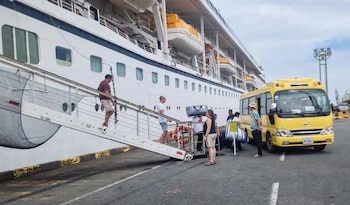 Pasajeros con equipaje bajan por una rampa desde un barco crucero hacia el muelle, donde espera un autobús amarillo