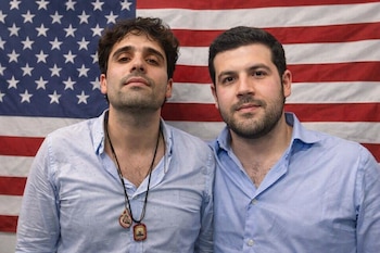 Primer plano de dos hombres con barba y camisas celestes, de pie frente a una bandera de Estados Unidos. Uno lleva dos collares con dijes.