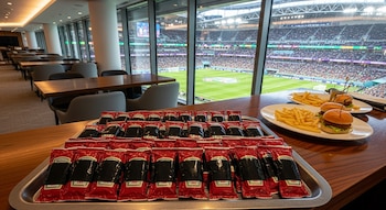 Una bandeja con muchos sobres de kétchup con cinta negra, dos platos con hamburguesas y papas fritas, un palco VIP y un estadio de fútbol lleno de gente.