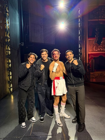 Quatro homens sorridentes no palco. O do meio está vestido como um boxeador ferido, com short branco, faixa na cabeça e sangue falso. As outras três roupas escuras.
