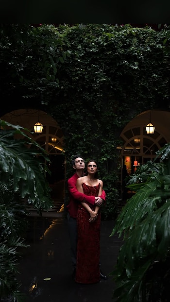 Hombre y mujer abrazados, ambos de pie y mirando hacia arriba, rodeados de abundante vegetación verde, plantas grandes, hiedra y faroles iluminados