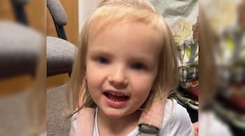 Una niña de cabello rubio con camiseta blanca sonríe y tiene los ojos difuminados.