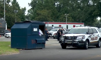Una patrulla de policía blanca y azul está estacionada frente a un contenedor de basura azul con oficiales y otros vehículos en el fondo