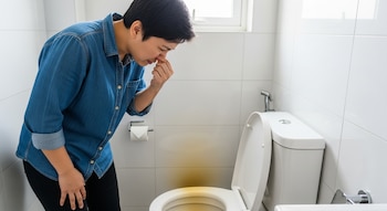 Una persona con camisa de mezclilla se inclina sobre un inodoro blanco en un baño. Se está tapando la nariz mientras una mancha amarilla rodea la taza del inodoro.