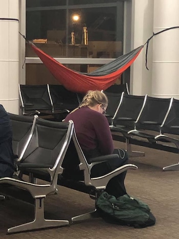 Una persona con trenzas y gafas sentada en una silla de aeropuerto. Una hamaca roja y gris cuelga entre dos pilares blancos frente a una ventana oscura
