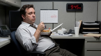 Un hombre de mediana edad con camisa y corbata come comida para llevar en un cubículo de oficina. Detrás, un reloj digital marca 9:45 PM y un calendario tiene "OVERTIME" circulado.