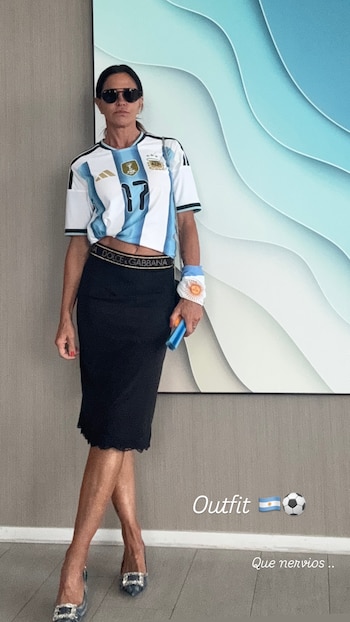 Una mujer de pie, con gafas de sol, camiseta de la selección argentina número 17, falda negra y zapatos de tacón, sostiene un objeto azul