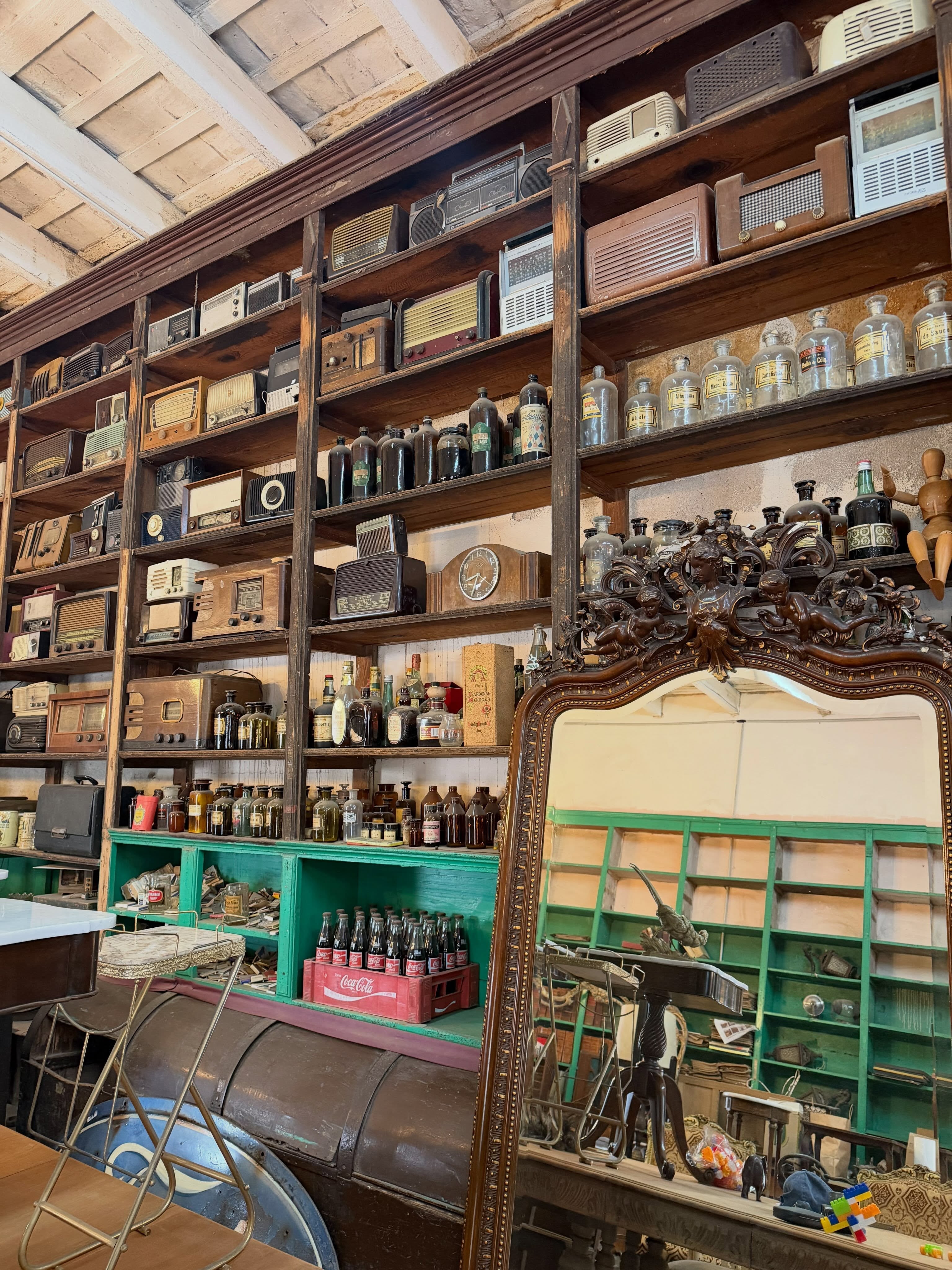 Una tienda de antigüedades exhibe repisas de madera repletas de radios antiguas, botellas de vidrio y un espejo tallado