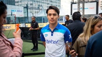 Un joven con camiseta deportiva blanca y azul permanece de pie en un espacio con césped artificial rodeado de personas con teléfonos.