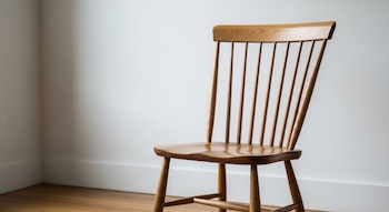 Primer plano de una silla Shaker de madera con respaldo de varillas verticales y asiento contorneado, situada contra una pared blanca y un suelo de madera.