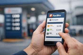 Manos sostienen un teléfono móvil con la aplicación Facilito abierta, que muestra precios de combustible. De fondo, una estación de servicio borrosa con surtidores.