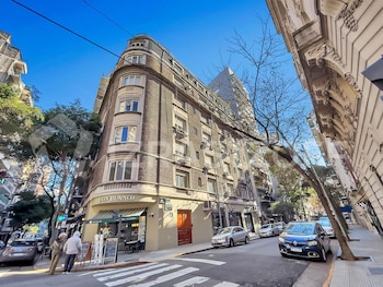 Edificio esquinero de ladrillo con un local comercial en la planta baja, una calle con automóviles estacionados, peatones, árboles y cielo azul